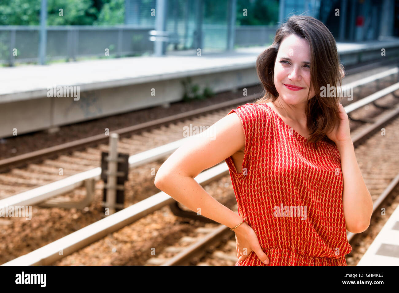 young woman in red dress waiting at platform of train station and smiles Stock Photo