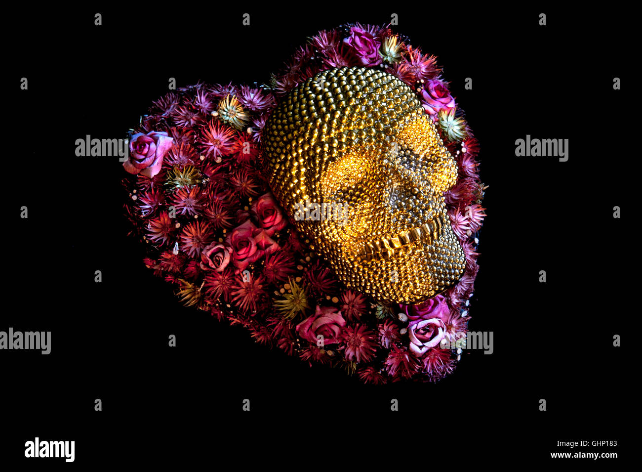 Concept image of smiling gold skull lying on heart shaped flower arrangement on black background Stock Photo