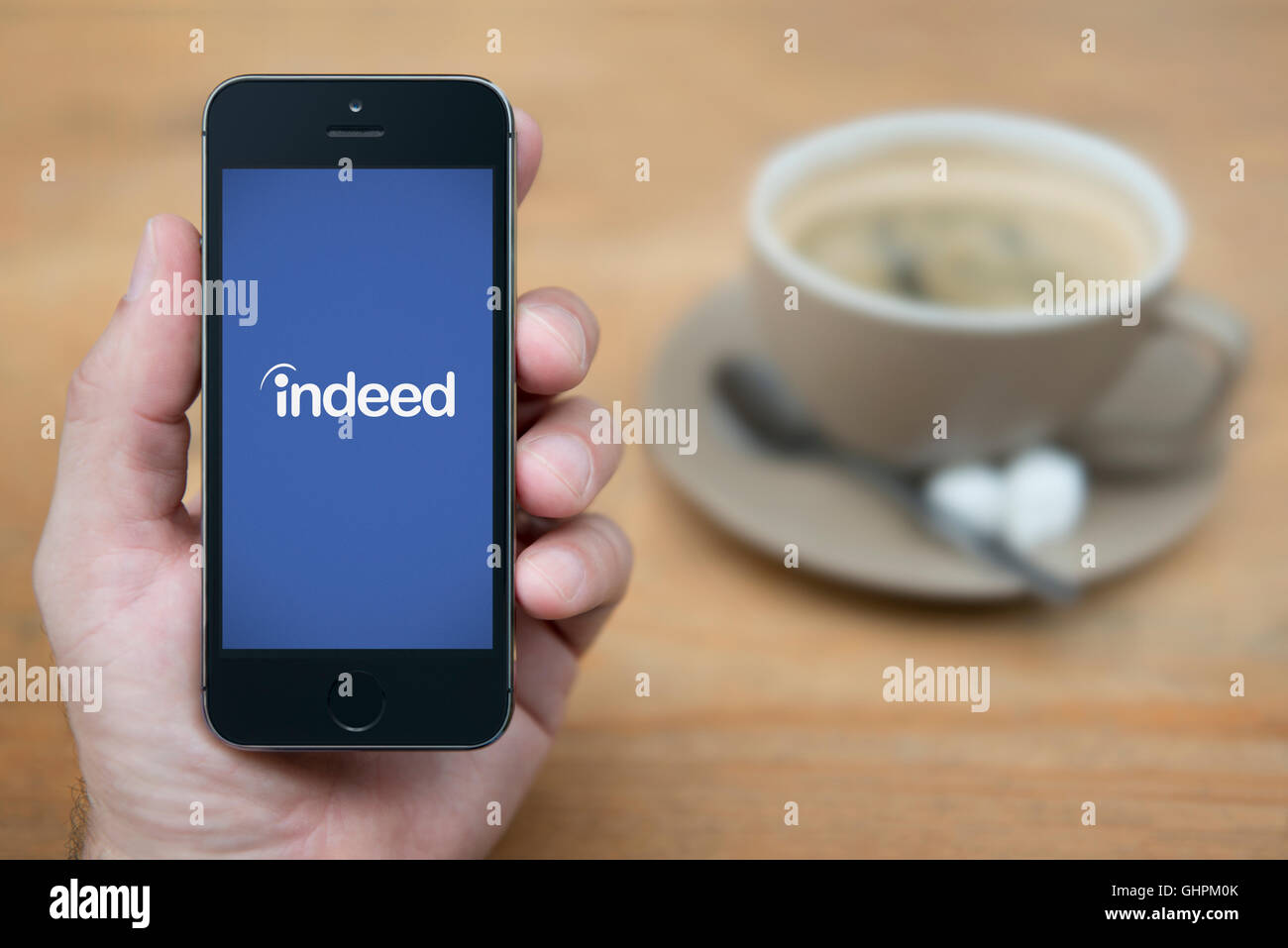A man looks at his iPhone which displays the Indeed logo, while sat with a cup of coffee (Editorial use only). Stock Photo