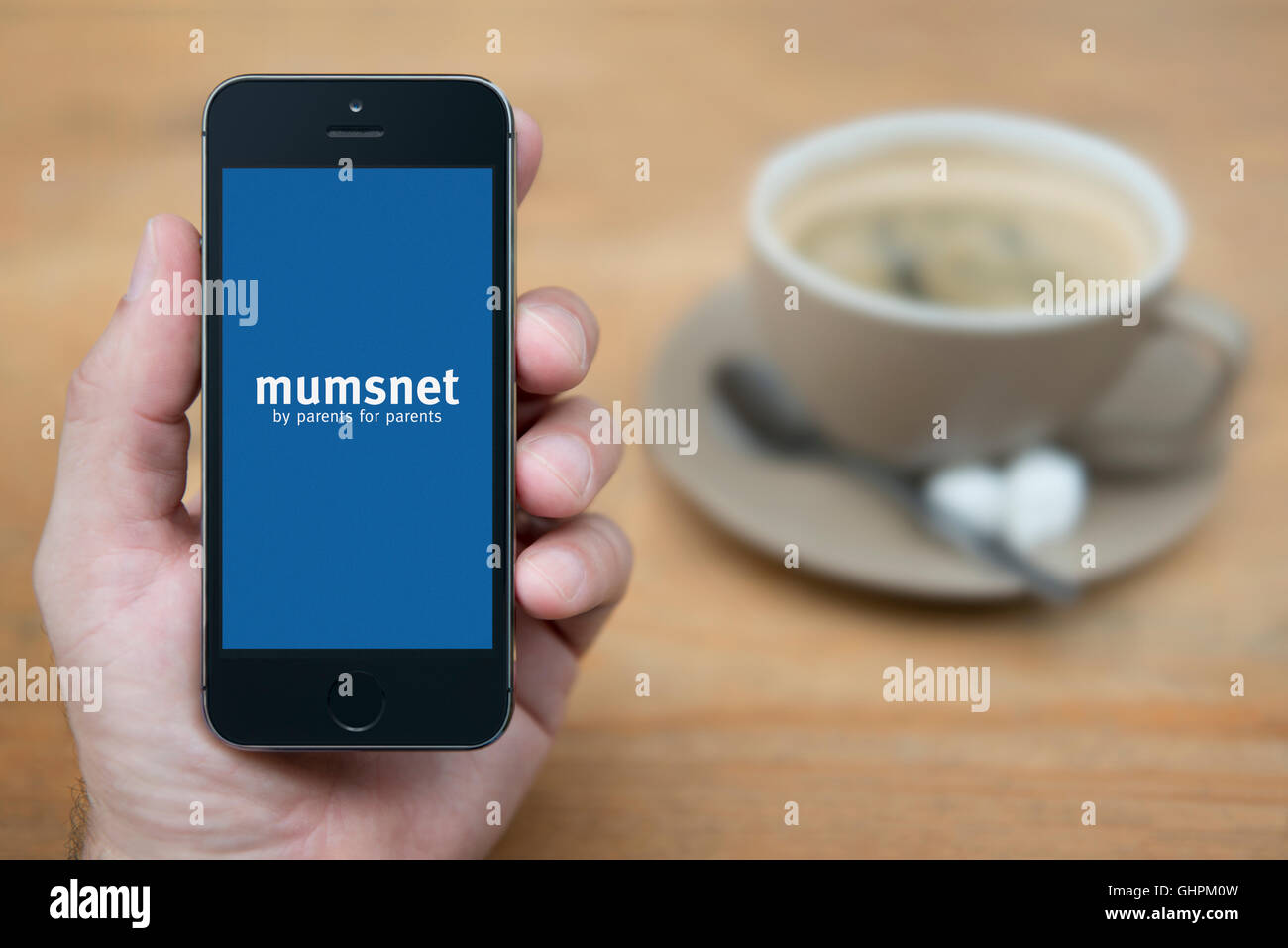 A man looks at his iPhone which displays the Mumsnet logo, while sat with a cup of coffee (Editorial use only). Stock Photo