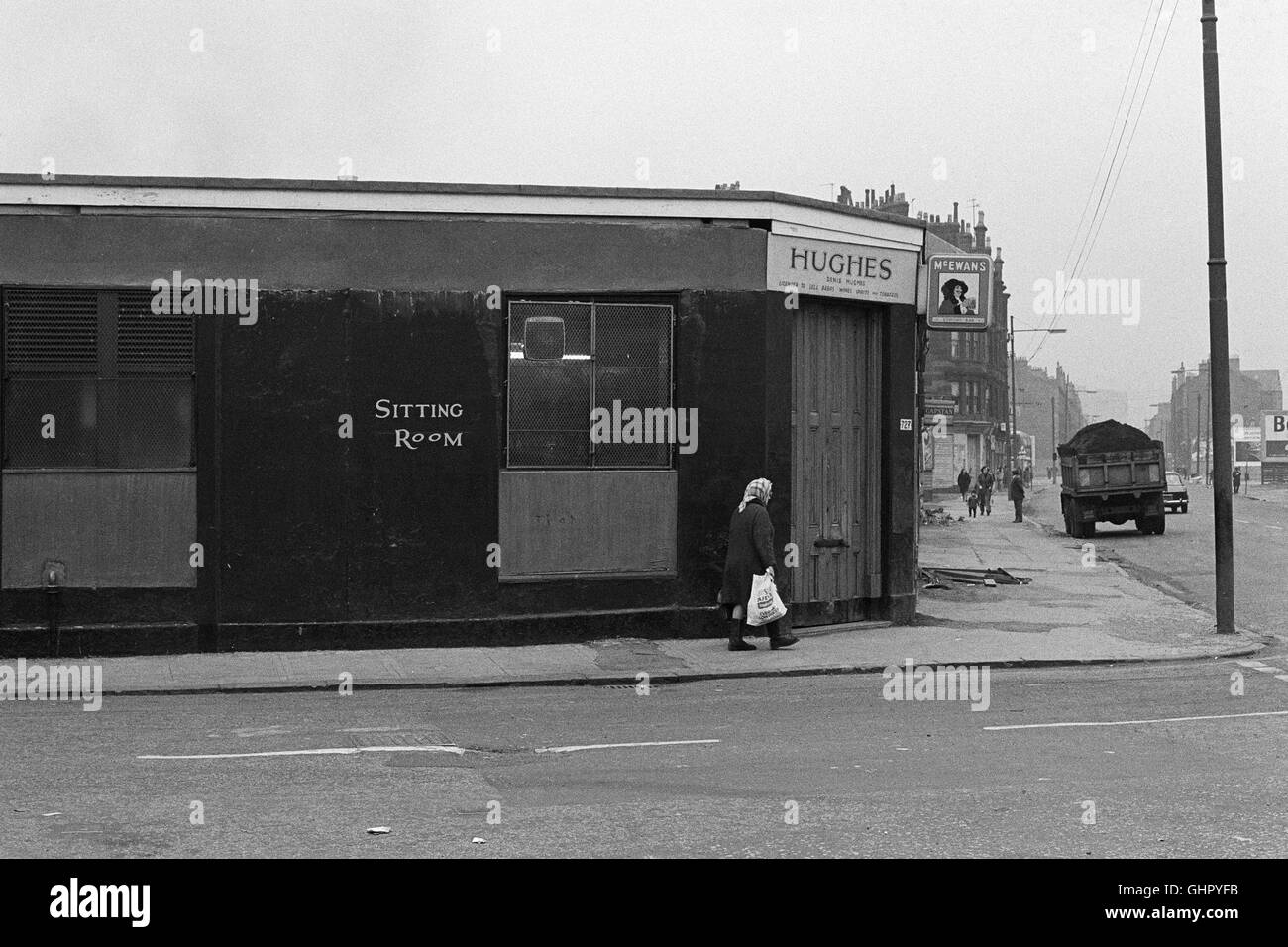 An old woman with a bag and headscarf walking on the pavement outside Hughes Public Bar in the East End Glasgow in 1971 approx. Stock Photo