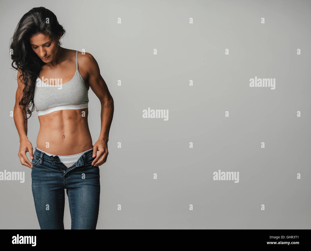 Woman standing with hands by her pockets looks downward while wearing unzipped jeans and halter top Stock Photo