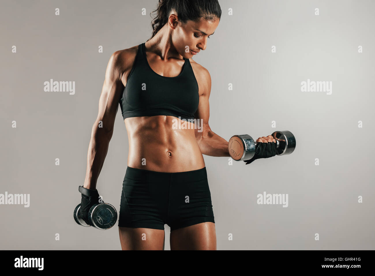 Single healthy athletic woman in black clothing with muscular body lifting chrome finish dumbbell weights over gray background w Stock Photo