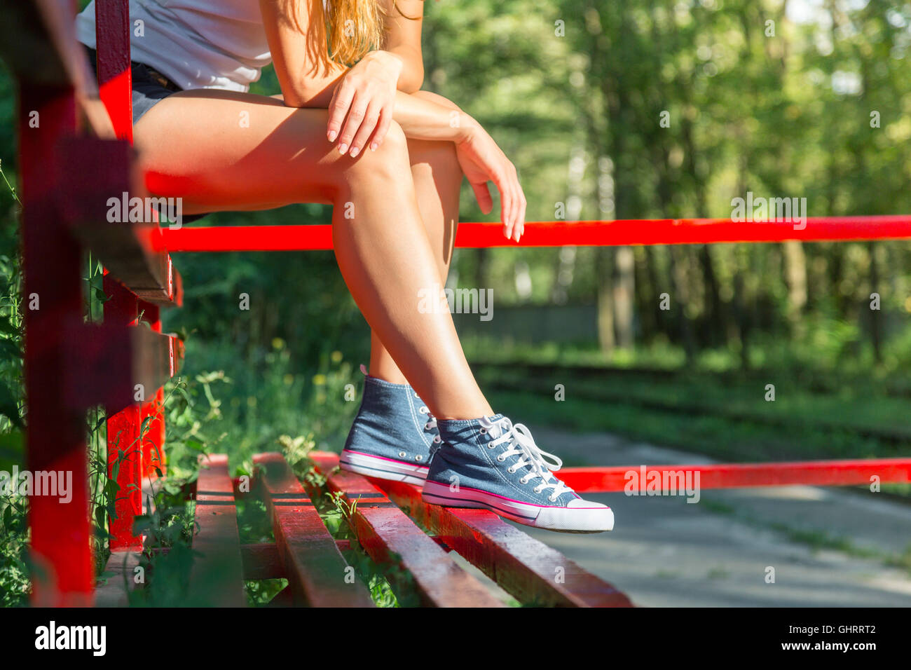 Young woman with beautiful sporty legs sitting on a red bus stop in jeans sneakers Stock Photo