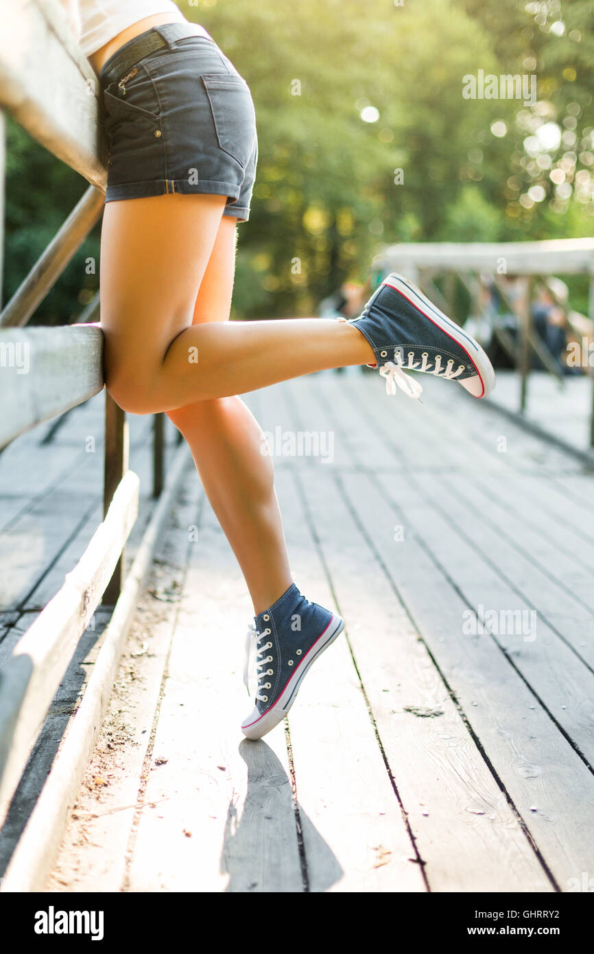Young woman with beautiful sporty legs leaning on a wooden bridge railing in jeans sneakers Stock Photo