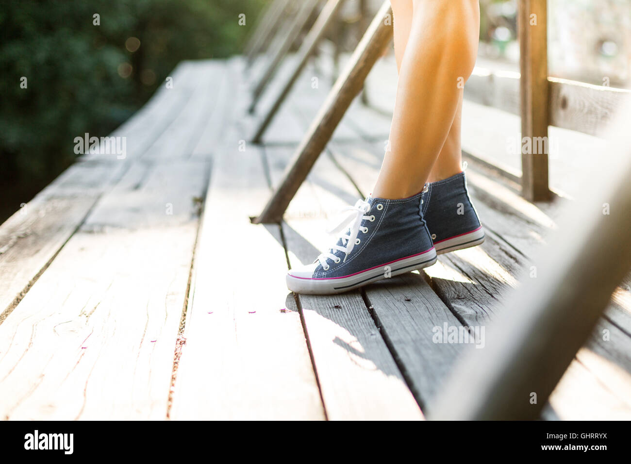 Young woman with beautiful sporty legs standing on a wooden bridge railing in jeans sneakers Stock Photo