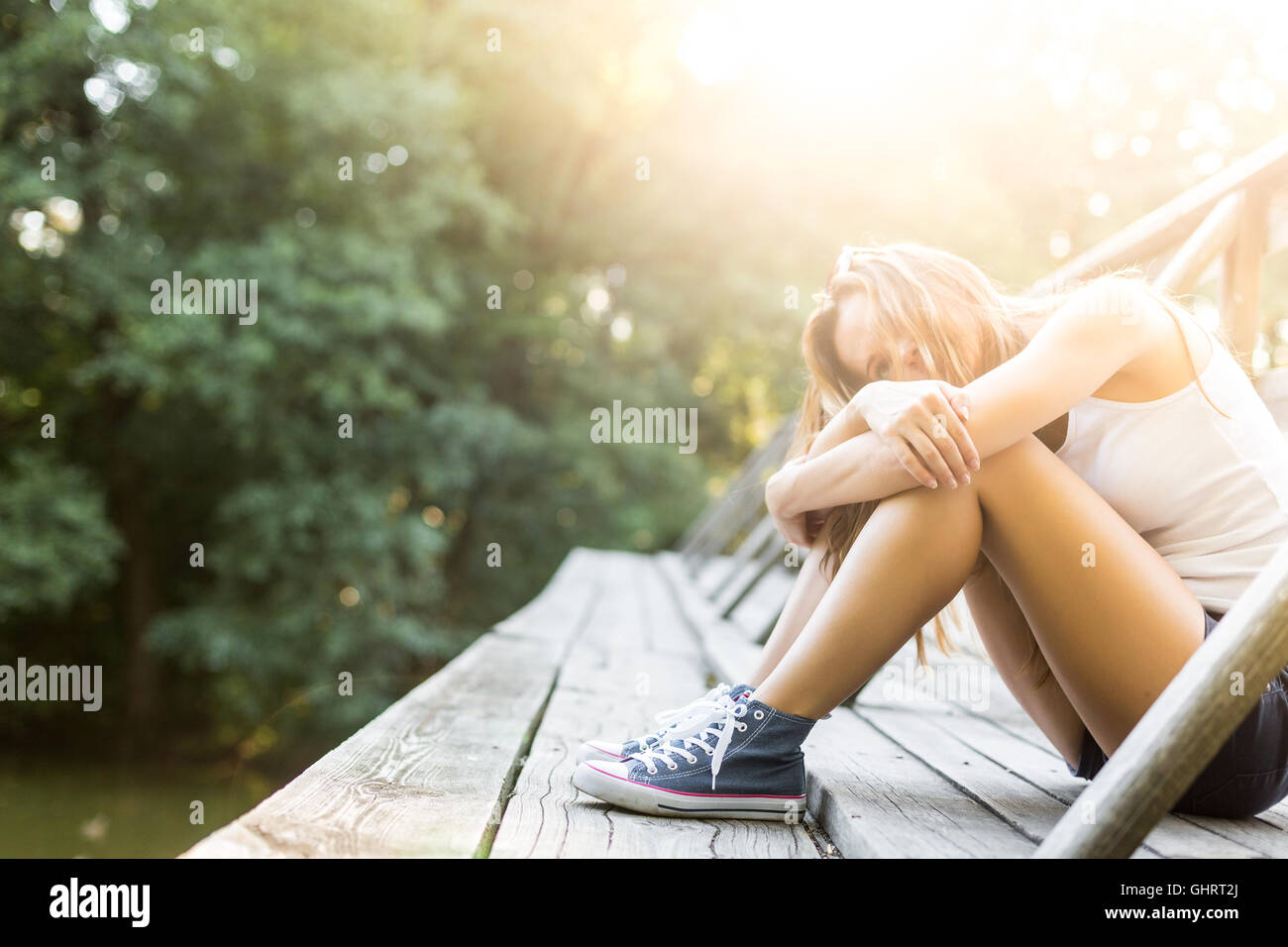 Young woman with beautiful sporty legs sitting on a wooden bridge railing in jeans sneakers Stock Photo