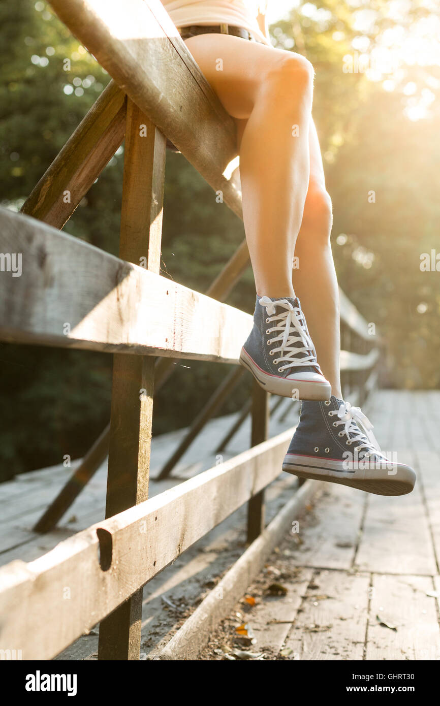 Young woman with beautiful sporty legs sitting on a wooden bridge railing in jeans sneakers Stock Photo