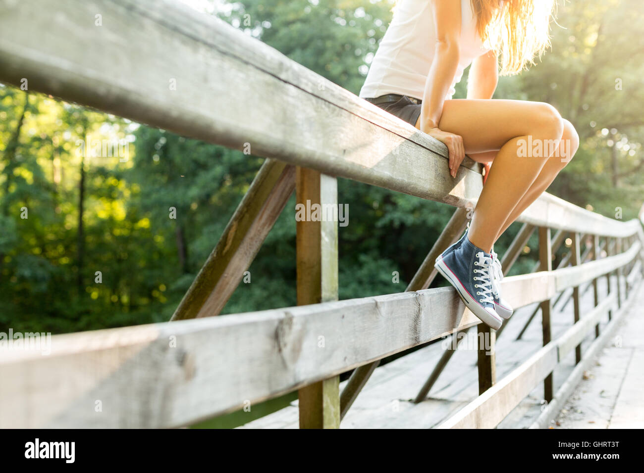 Young woman with beautiful sporty legs sitting on a wooden bridge railing in jeans sneakers Stock Photo
