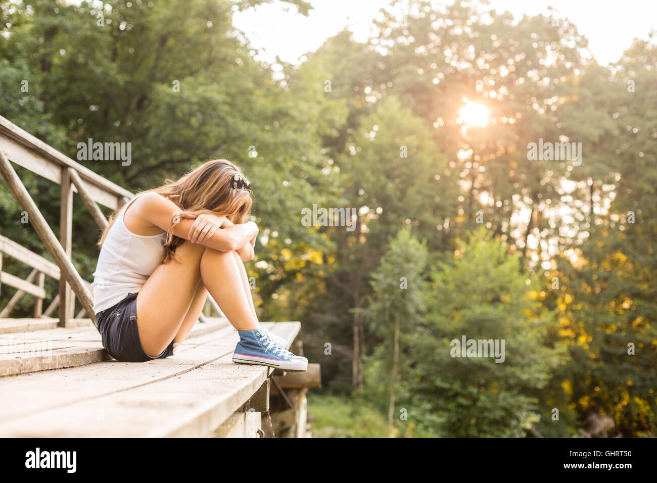 Young sad woman with beautiful sporty legs sitting on a wooden bridge railing in jeans sneakers Stock Photo