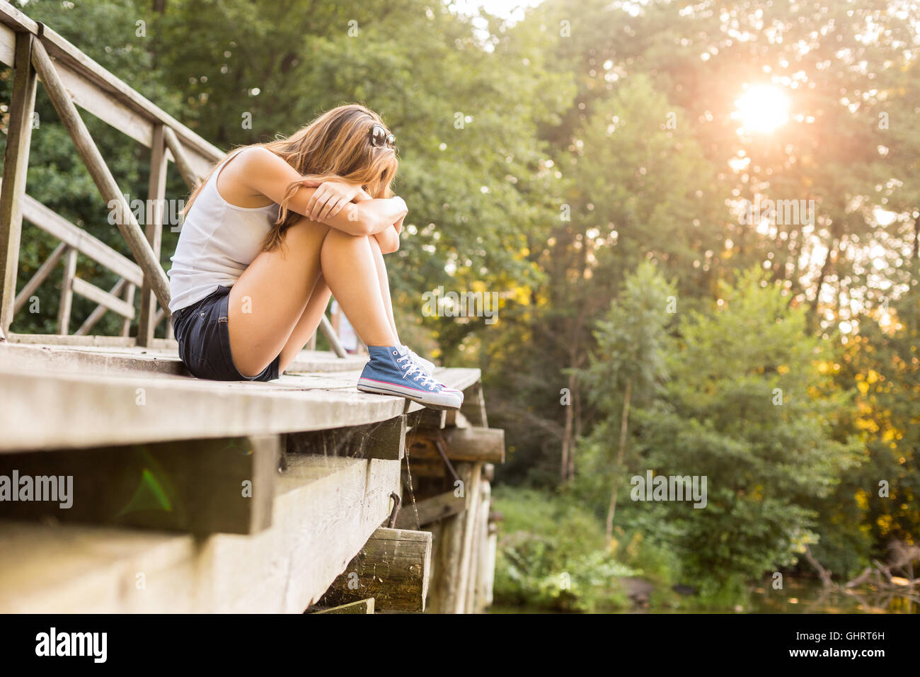 Young sad woman with beautiful sporty legs sitting on a wooden bridge railing in jeans sneakers Stock Photo
