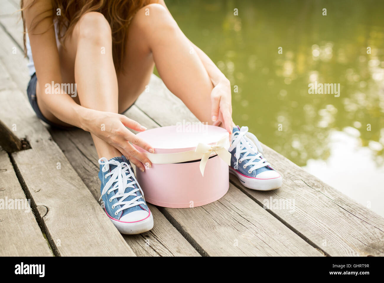 Young woman with beautiful sporty legs in jeans sneakers  sitting on a wood  opening pink gift box Stock Photo