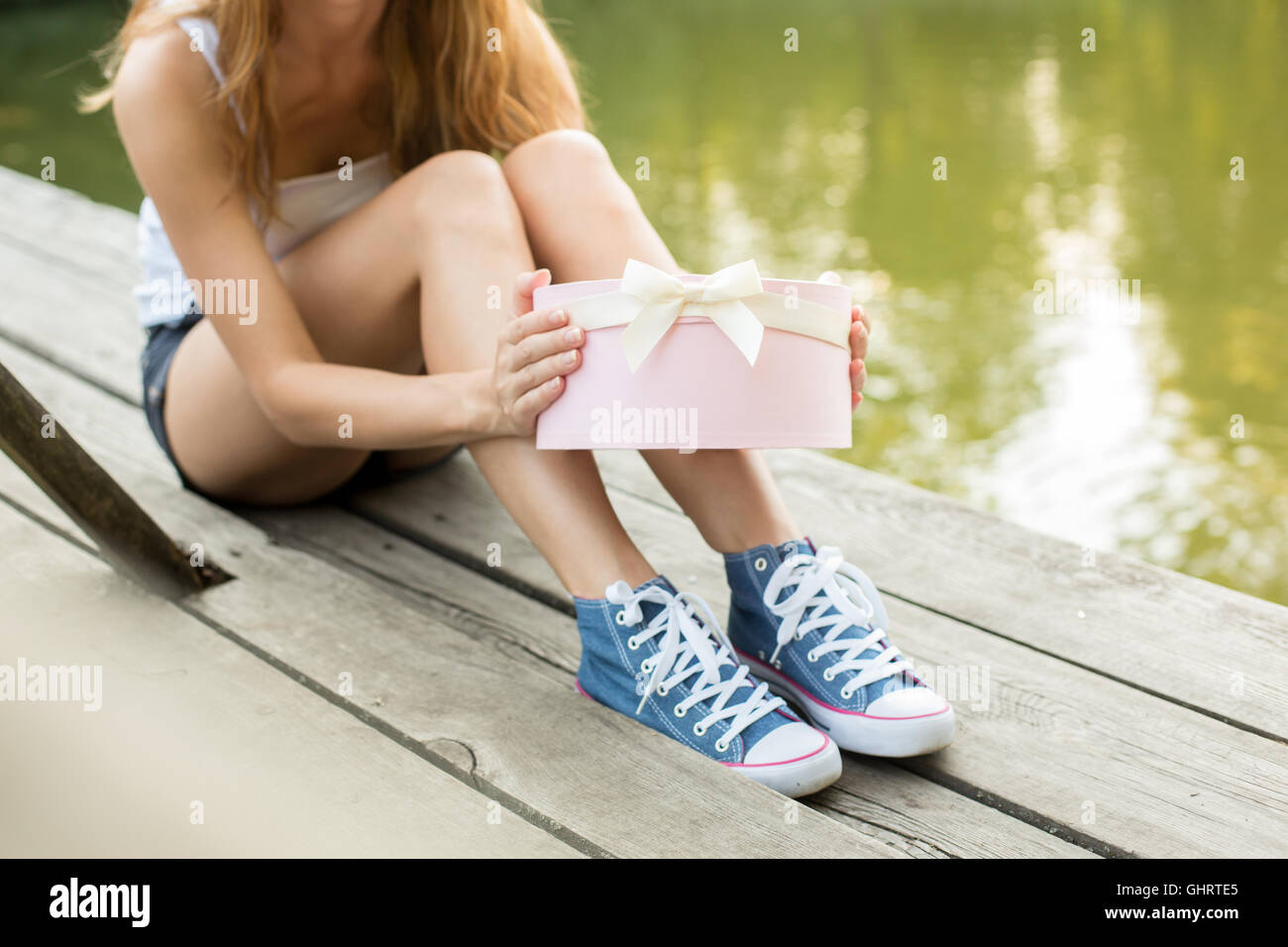 Young woman with beautiful sporty legs in jeans sneakers  sitting on a wood  opening pink gift box Stock Photo