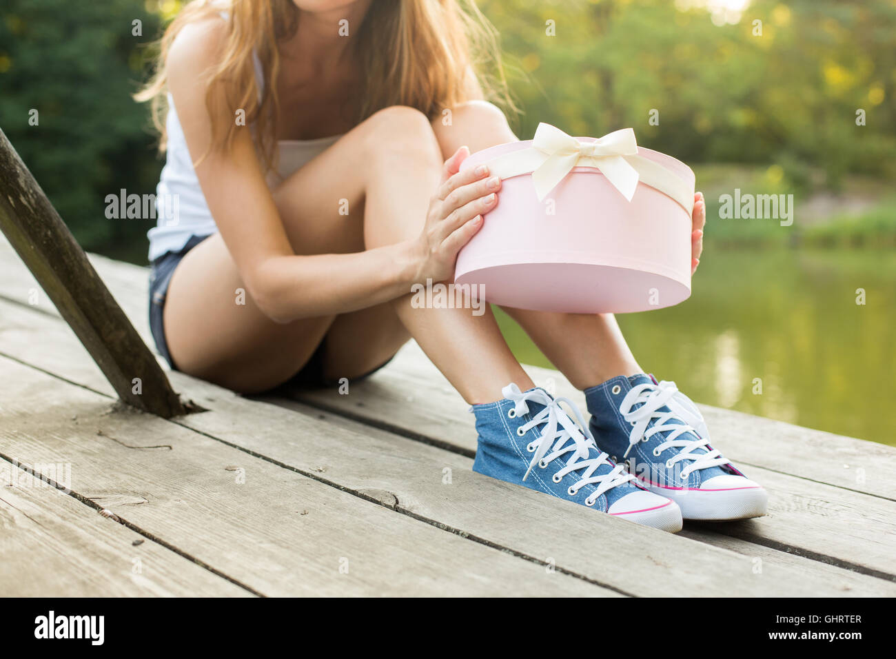 Young woman with beautiful sporty legs in jeans sneakers  sitting on a wood  opening pink gift box Stock Photo