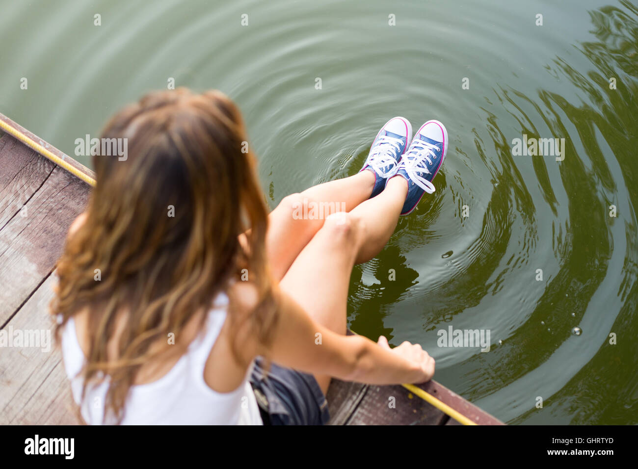 Young woman wearing jeans sneakers sitting in a river bank hanging her  legs over the water Stock Photo