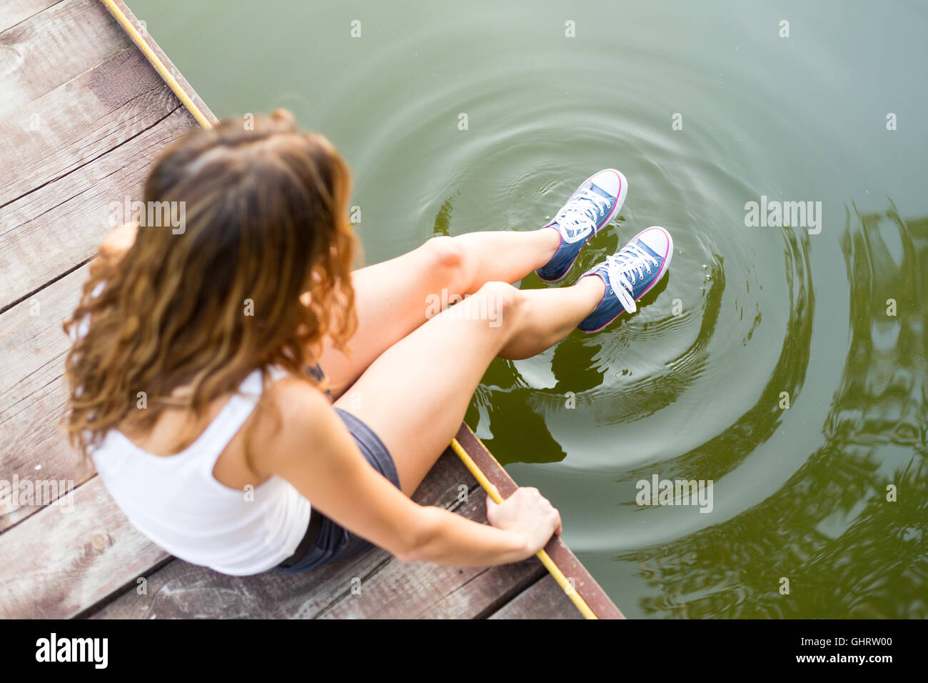 Young woman wearing jeans sneakers sitting in a river bank hanging her  legs over the water Stock Photo