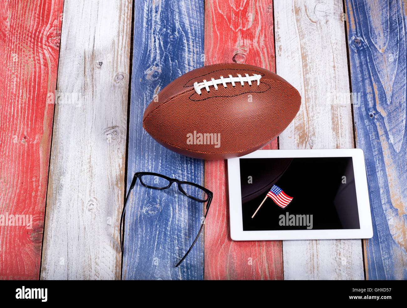 Overhead view of mobile computer, reading glasses, and American football on red, white and blue rustic wooden boards. Stock Photo