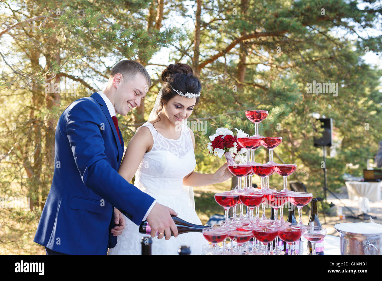 Newlyweds are pouring vine in the pyramid of glasses at the open air wedding banquet Stock Photo