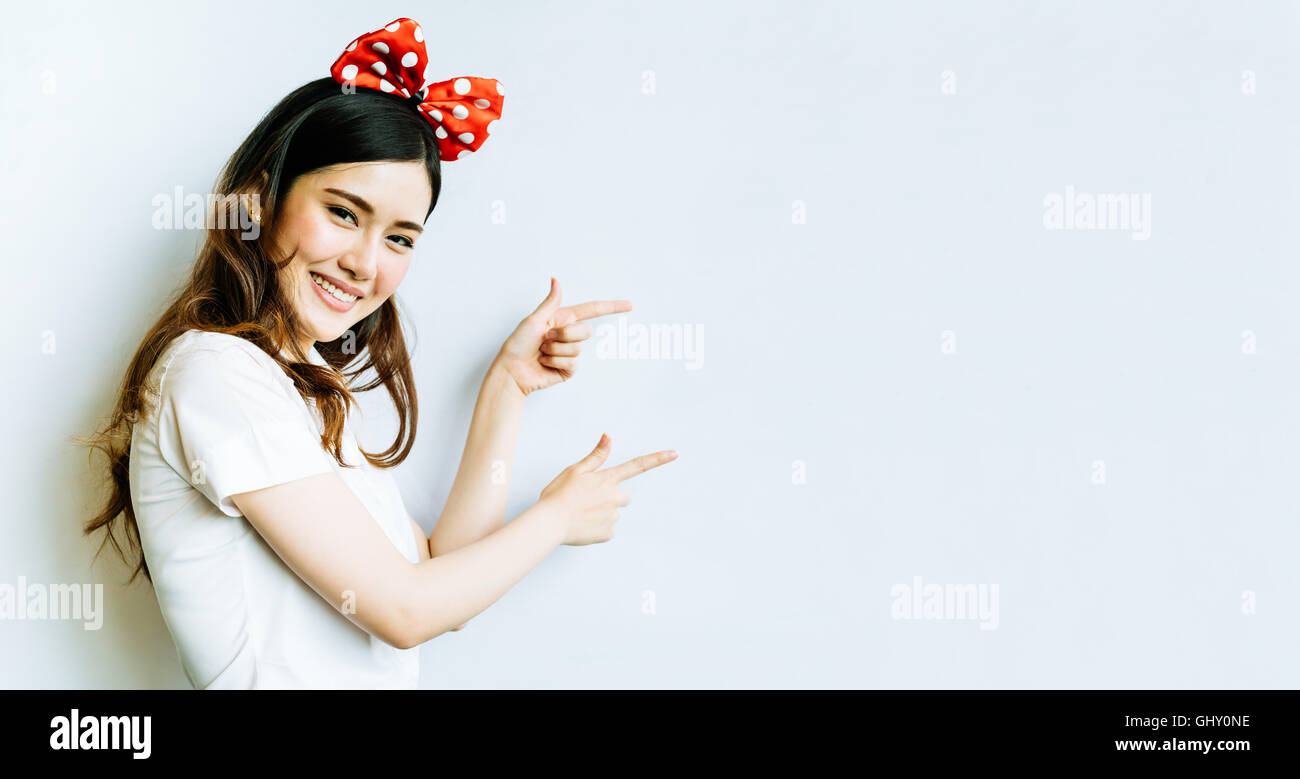 Beautiful asian university or college student woman wearing funny bow headband, pointing at copy space on whiteboard background Stock Photo