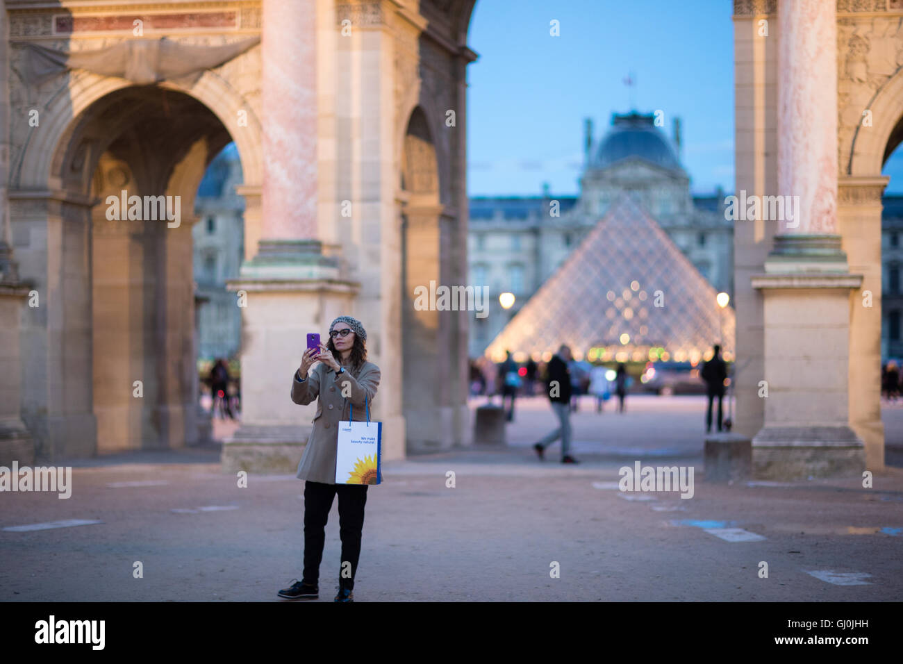 a woman taking a selfie at the Arc de Triomphe du Carrousel & Palais du Louvre at dusk, Paris, France Stock Photo