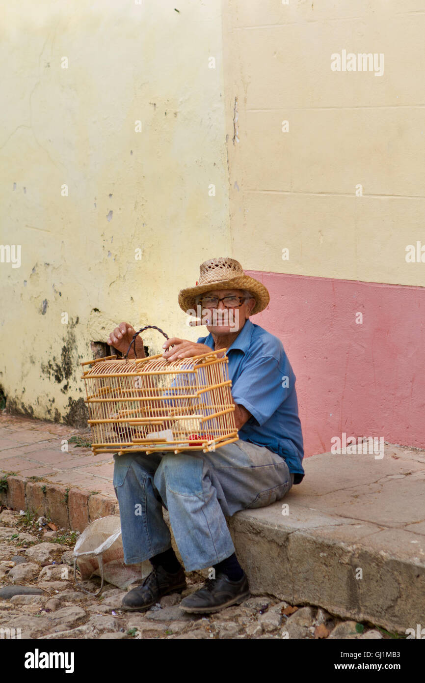 Old Man with birds in cage and cigar, sitting on pavement in Trinidad, Cuba, 2013 Stock Photo