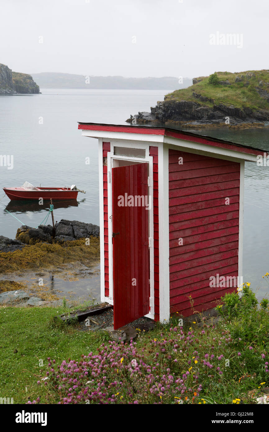 A red outhouse at Bay Roberts in Newfoundland and Labrador, Canada. A boat is moored just off the shore, in the water of Concept Stock Photo