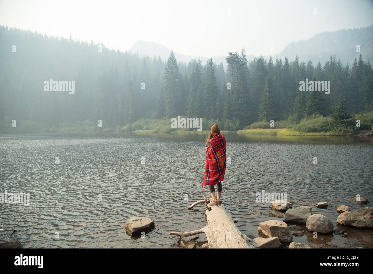 Woman wrapped in tartan blanket looking out over misty lake, Mount Hood National Forest, Oregon, USA Stock Photo