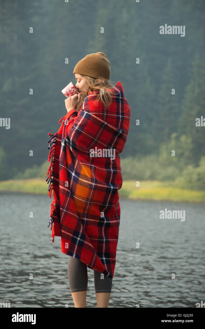 Woman wrapped in tartan blanket drinking coffee by lake, Mount Hood National Forest, Oregon, USA Stock Photo