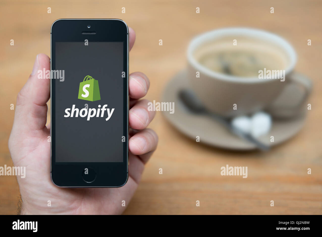 A man looks at his iPhone which displays the Shopify logo, while sat with a cup of coffee (Editorial use only). Stock Photo
