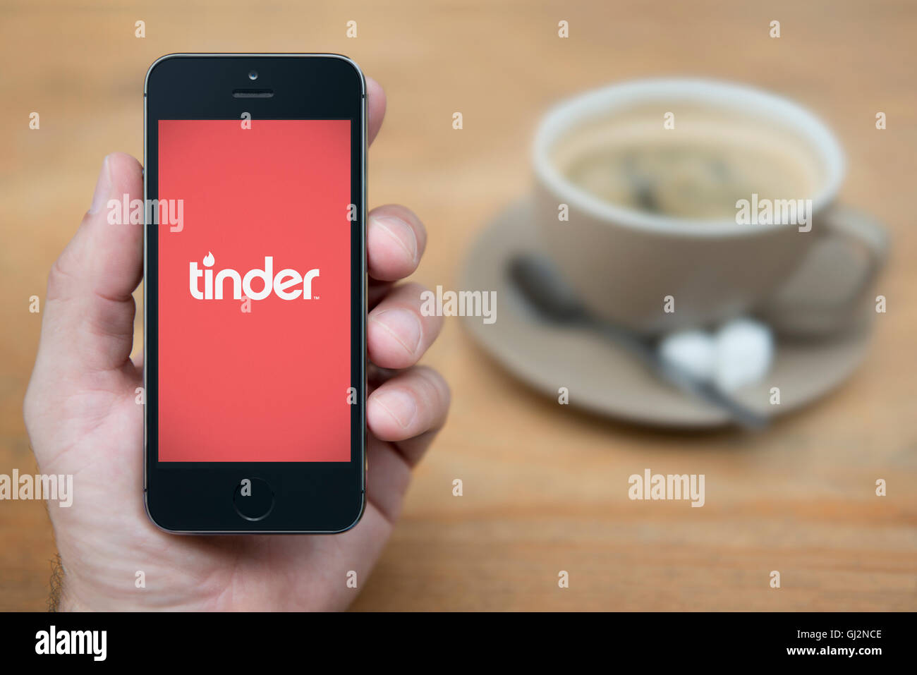 A man looks at his iPhone which displays the Tinder logo, while sat with a cup of coffee (Editorial use only). Stock Photo