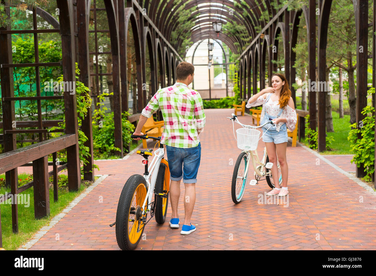 Meeting of a girl and guy in green and red plaid shirt with bikes in archway in the beautiful  park Stock Photo