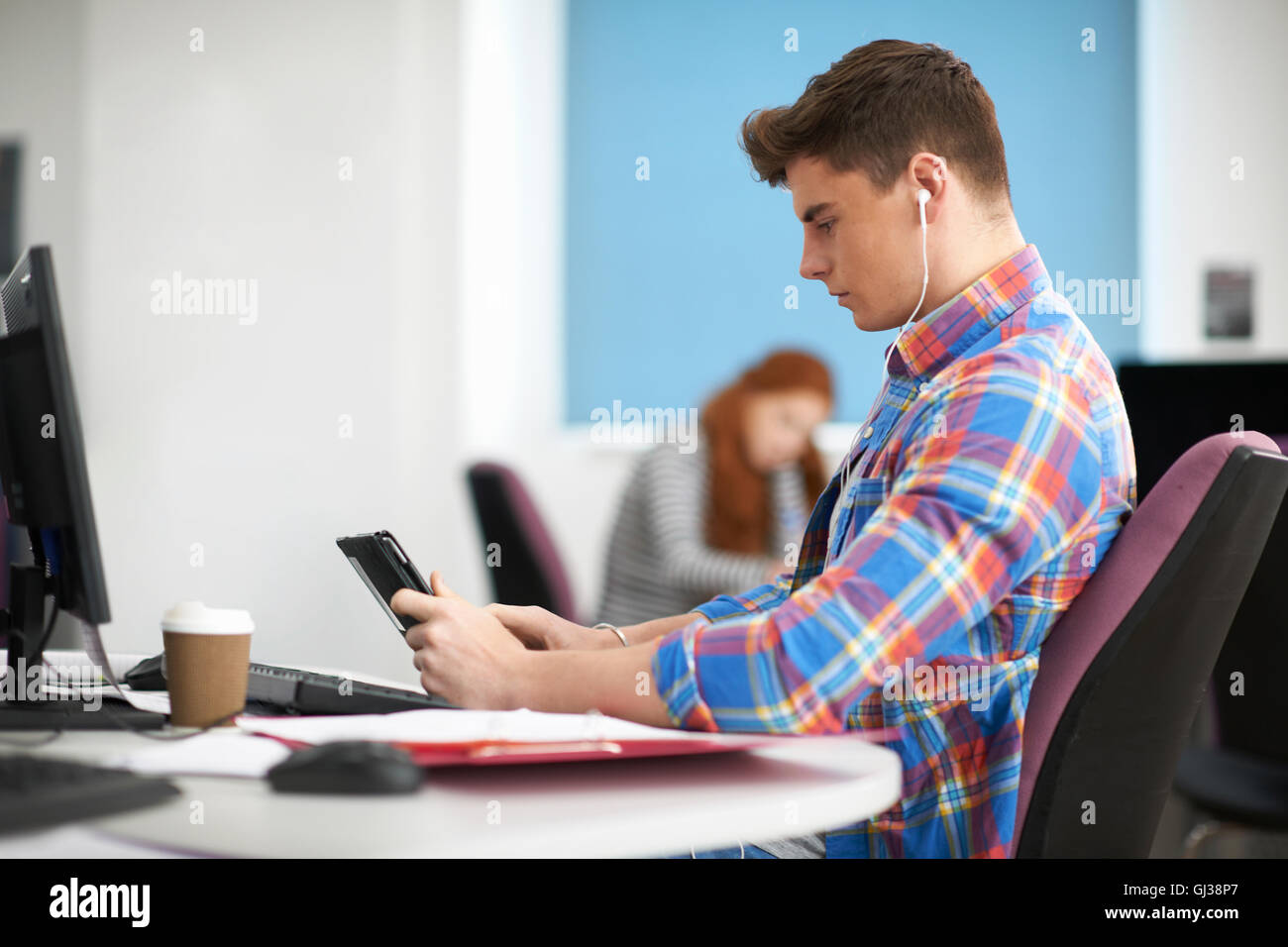 Young Male College Student At Computer Desk Looking At Smartphone