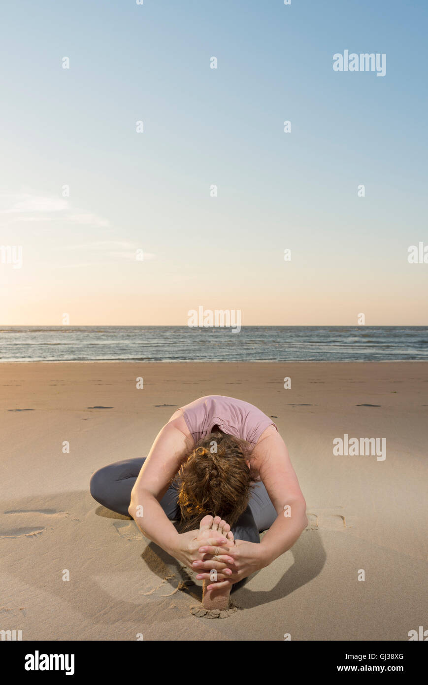 Mature woman practising yoga on a beach at sunset, touching toes Stock Photo