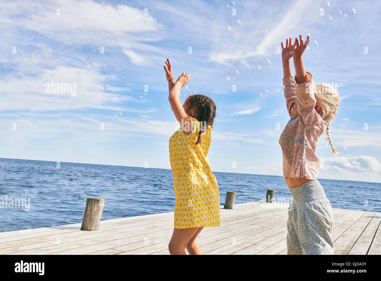 Two young friends playing on wooden pier, reaching for bubbles Stock Photo