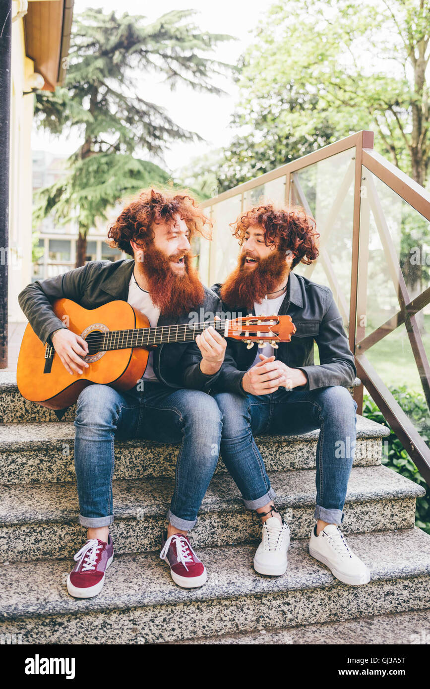Young male hipster twins with red beards sitting on stairway playing