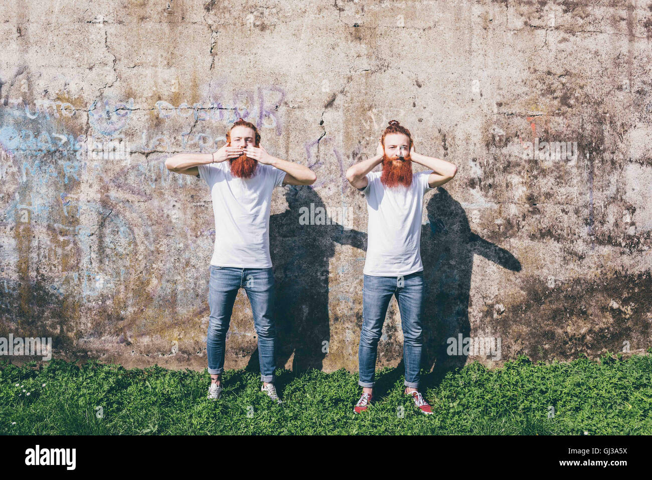 Portrait of young male hipster twins with red beards standing in front of wall with hands covering mouth and ears Stock Photo