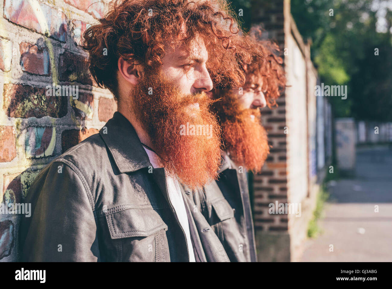 Young male hipster twins with red hair and beards leaning against brick wall Stock Photo