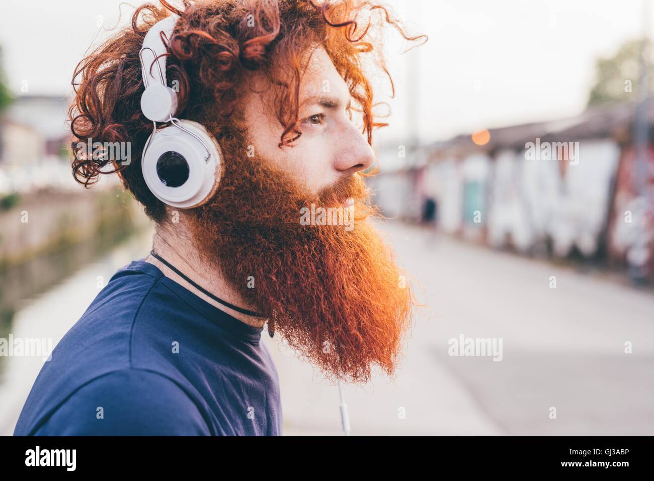 Young male hipster with red hair and beard listening to headphones in city Stock Photo