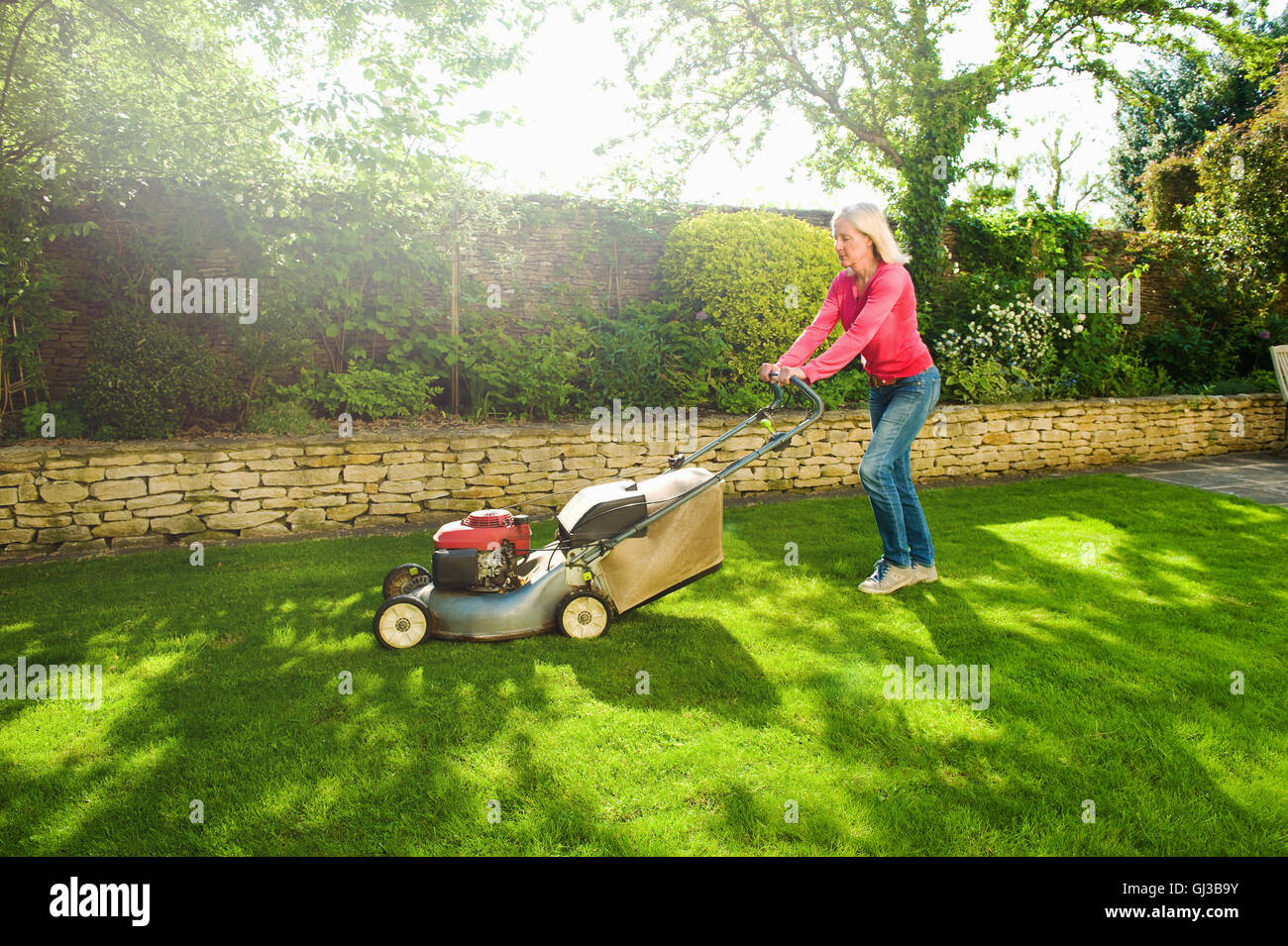 Mature woman mowing sunlit garden lawn with lawn mower Stock Photo