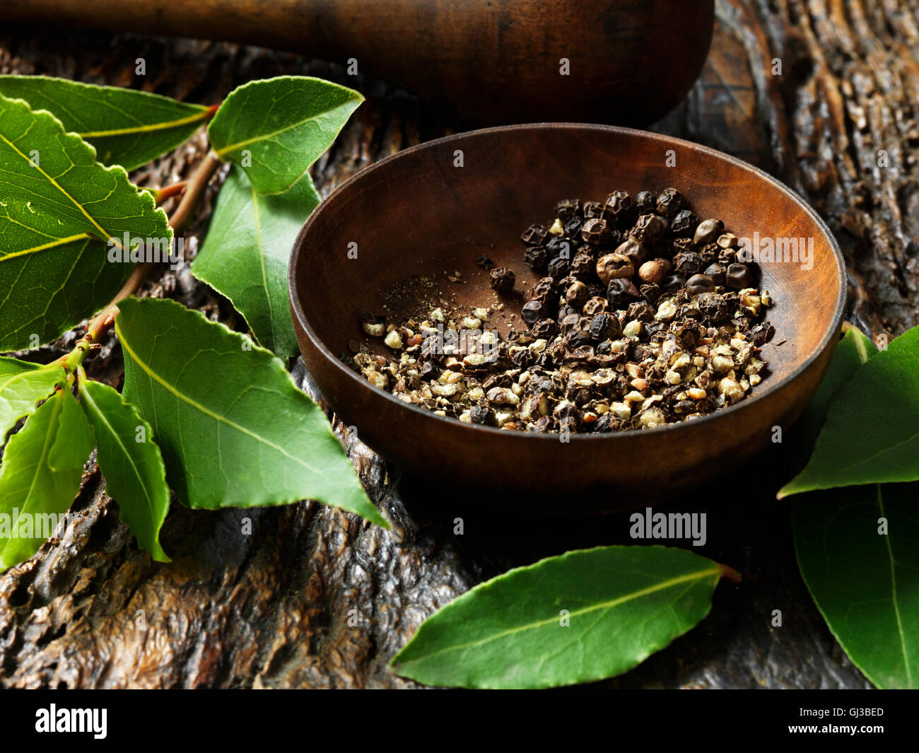 Crushed peppercorns in bowl, bay leaves on rustic wooden surface Stock Photo
