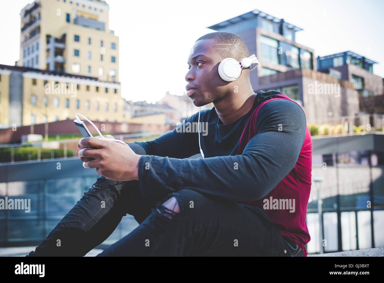 Young man outdoors, sitting on wall, holding smartphone, wearing headphones Stock Photo