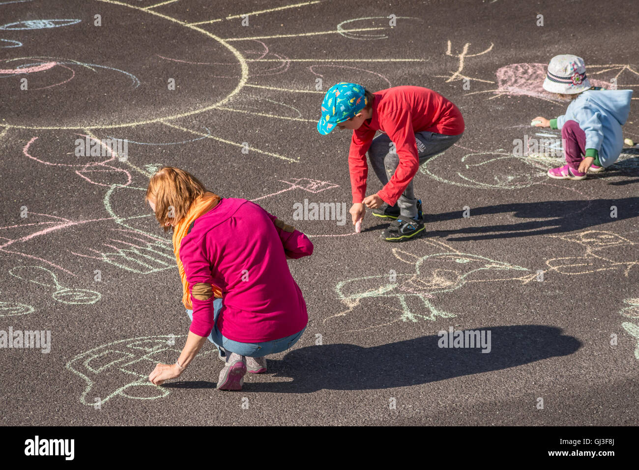 Caucasian mother playing with 2 two children as drawing figures with chalk on asphalt. Family outdoor activity. Stock Photo