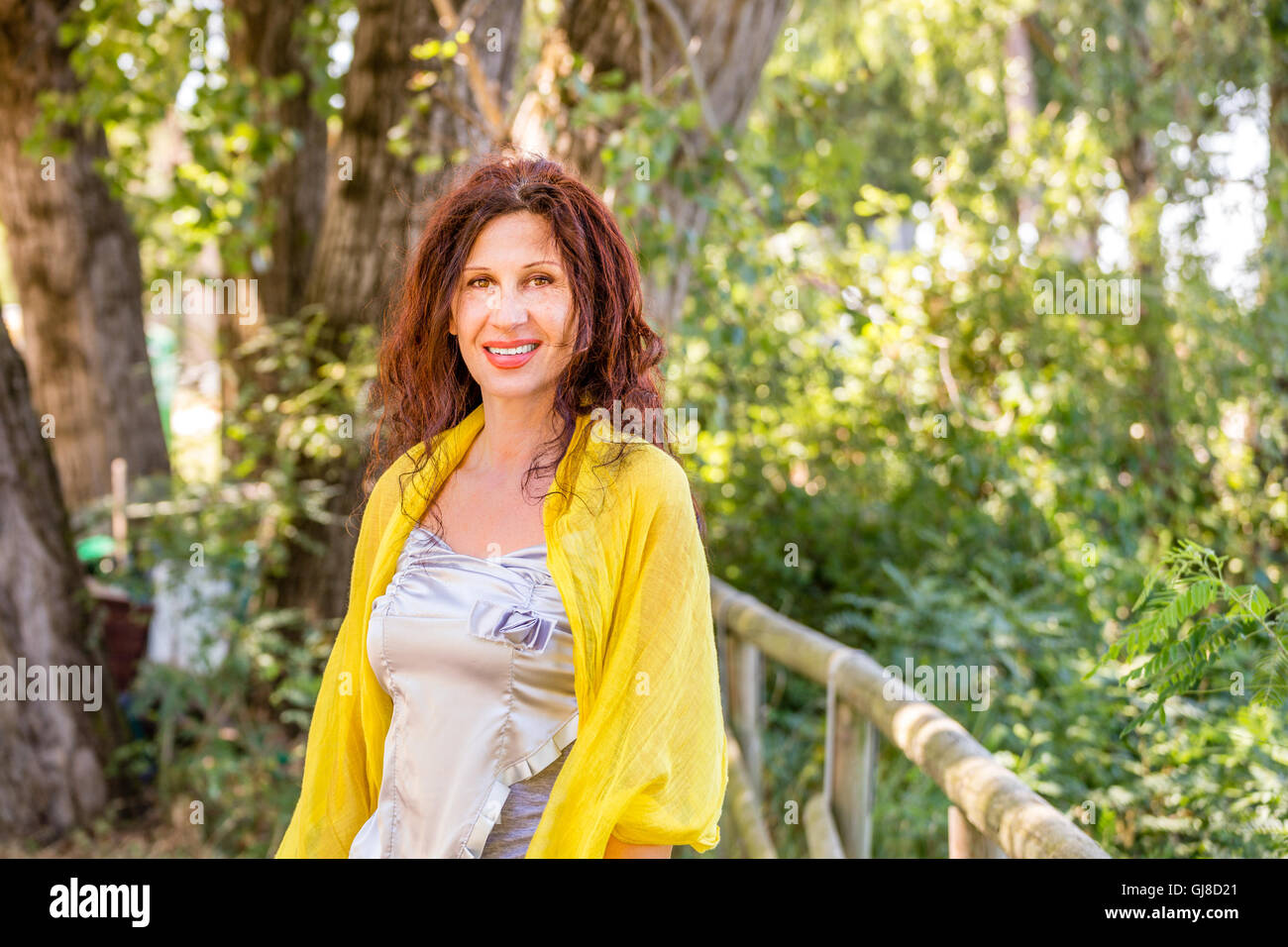 elegant and glamorous mature woman with yellow shawl and silver top is smiling in a green park Stock Photo