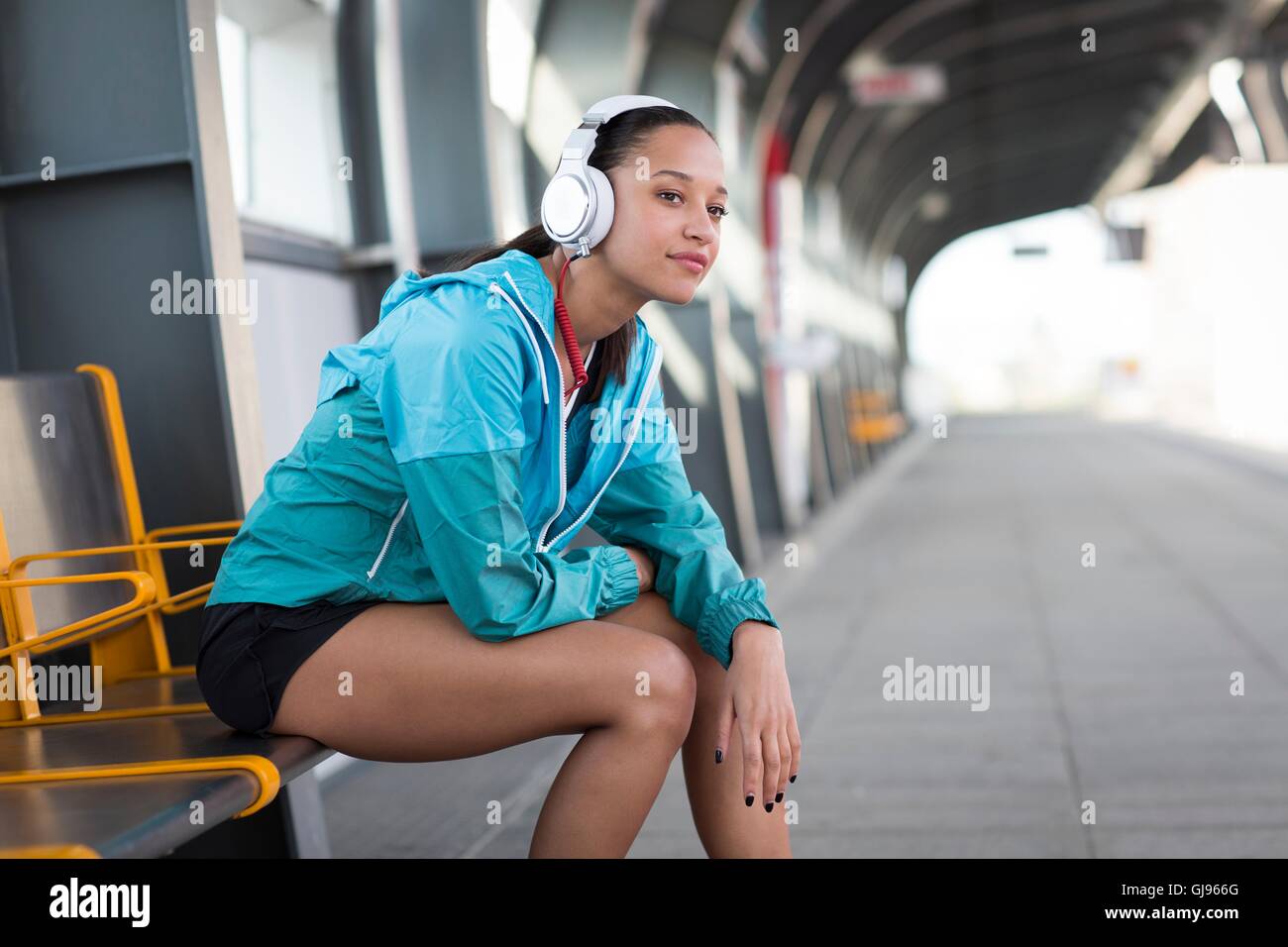 MODEL RELEASED. Young woman sitting on railway platform wearing headphones. Stock Photo