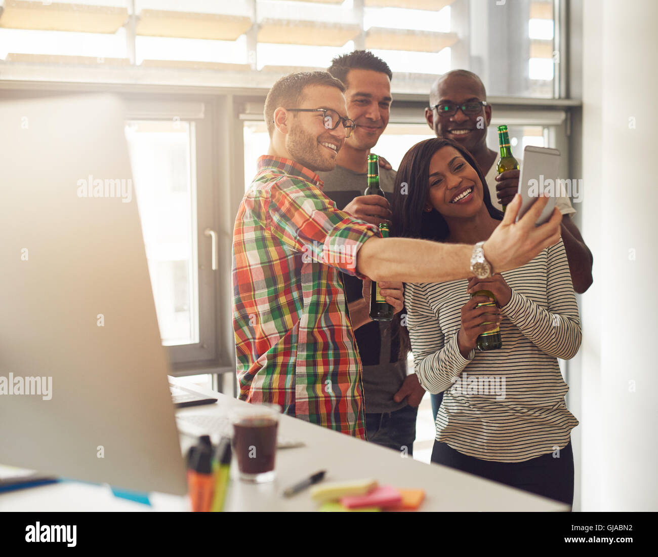 Diverse group of four happy young workers celebrating with beer and taking pictures with camera phone near desk with computer Stock Photo