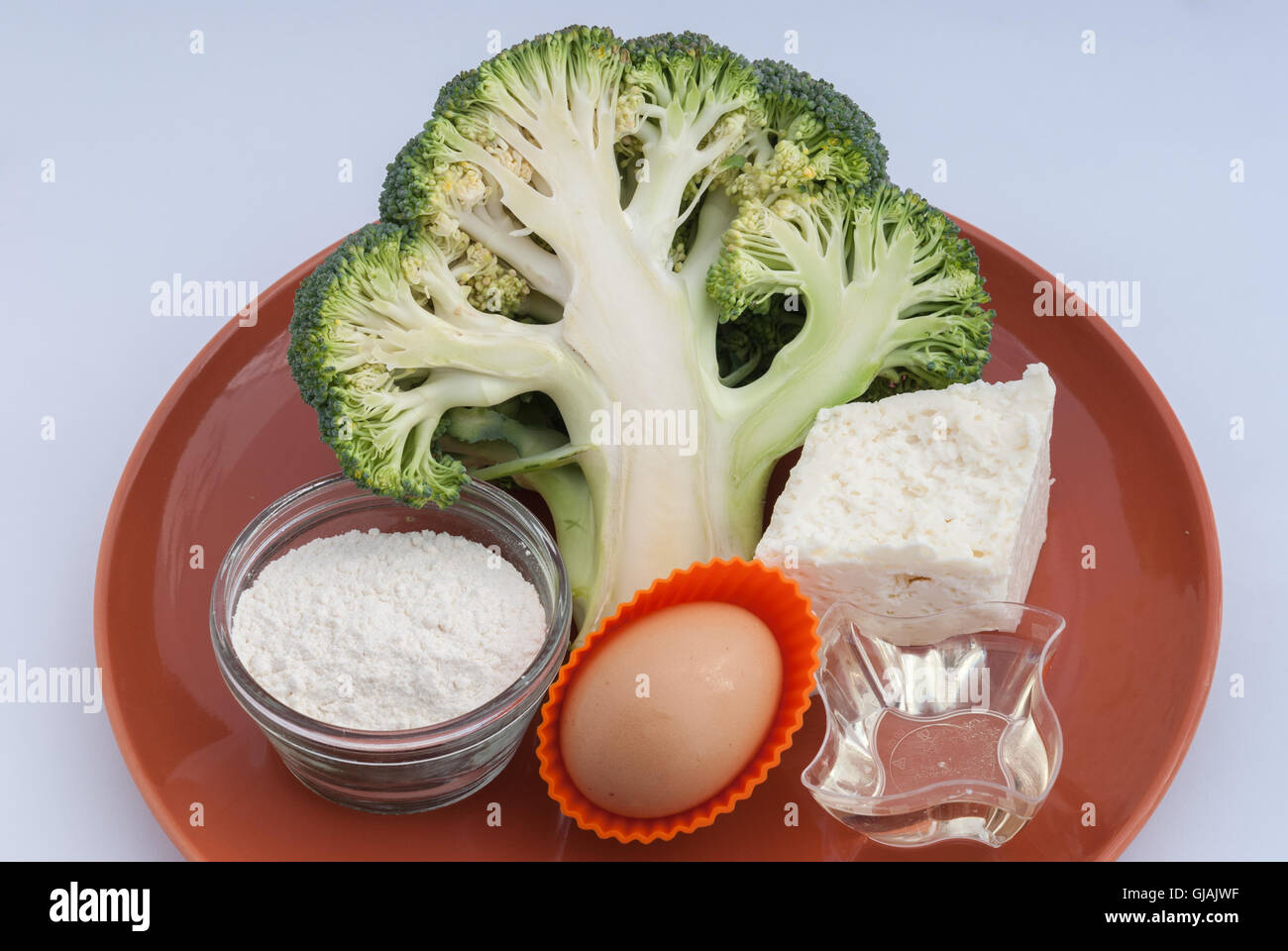 Cooking ingredients: fresh broccoli, brinza cheese, egg, sunflower oil and flour on a brown plate ready for cooking closeup. Stock Photo