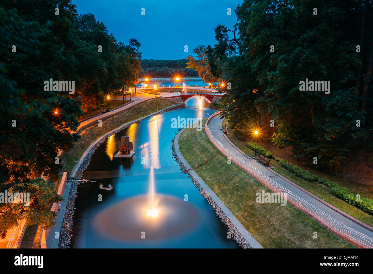 The Scenic View Of Park Watercourse Channel Flowing Into The River Through Stone Bridge. Lighted Walkways, Greenwood Along The W Stock Photo