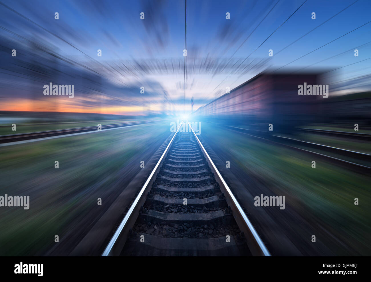 Railway station with cargo wagons in motion at sunset. Railroad with motion blur effect. Railway platform at dusk. Stock Photo