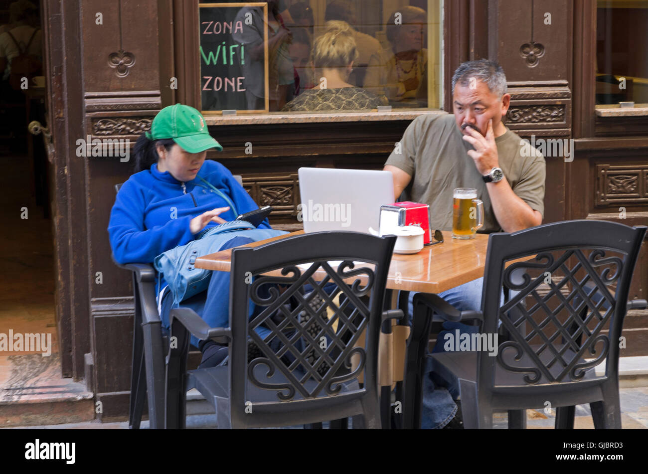 Older asian man and teen girl check internet devices at sidewalk café table. Stock Photo