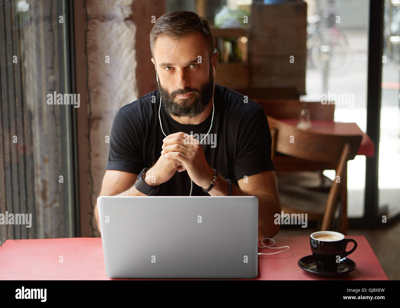 Handsome Young Bearded Businessman Wearing Black Tshirt Working Laptop Urban Cafe.Man Sitting Wood Table Cup Coffee Listening Music.Coworking Process Business Startup.Blurred Background. Stock Photo